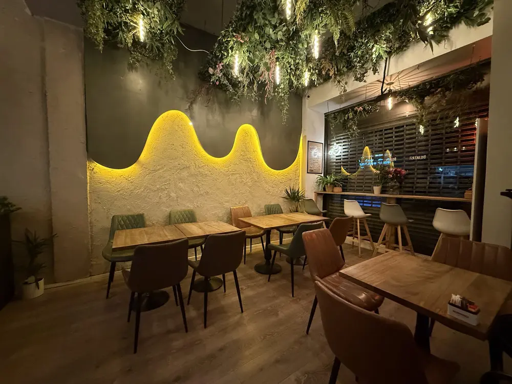 Love Is at night — a wavy plaster wall lit in warm gold, hanging greenery overhead and wooden tables set for brunch